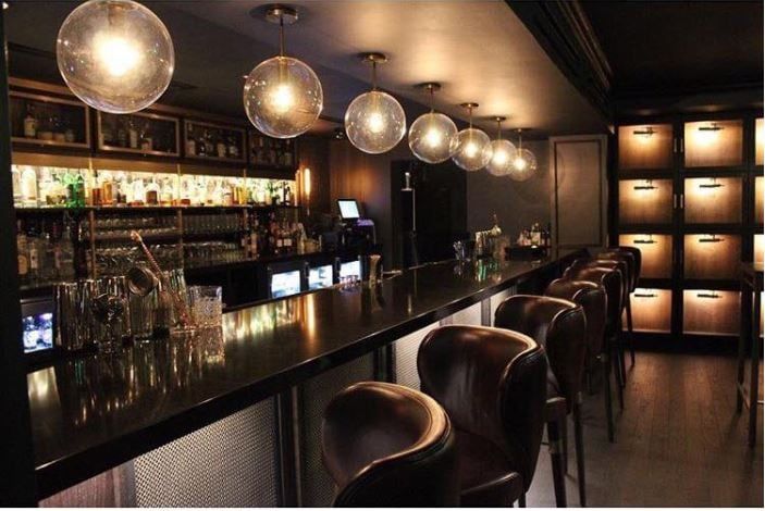 PS bar with shelves filled with bottles and circular lights hanging above the counter. There are empty chairs in front.