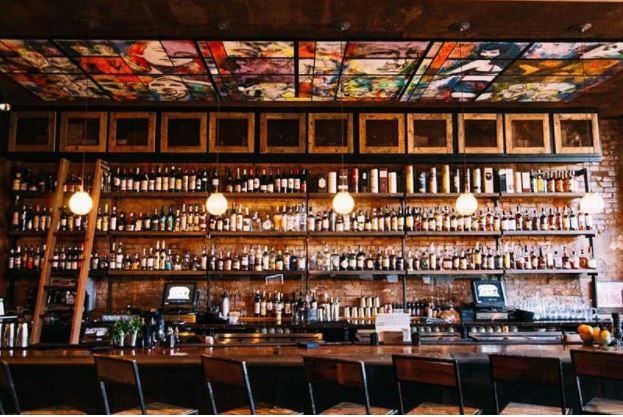Well-lit bar, a line of chairs in front of the counter with wooden shelves behind the counter filled with spirits.