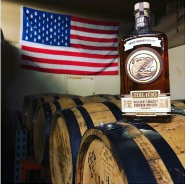 A bottle of whiskey placed on top of whiskey barrels with the American flag hanging on the wall behind it.