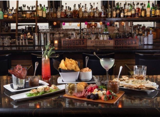 Bar counter filled with dishes, cocktails, and whiskeys on top. The shelves behind are filled with bottled drinks.